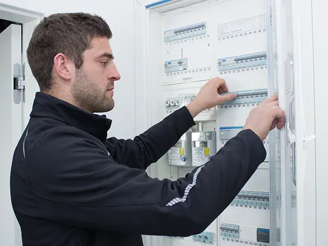 Mitarbeiter STREIF prüft Elektroverteiler mit Sicherungen in hellem Technikraum aus Seitenansicht