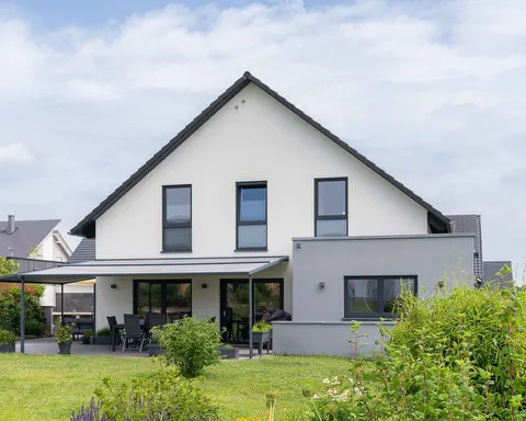 Fertighaus Einfamilienhaus mit weißer Fassade, Satteldach, grauem Anbau und überdachter Terrasse im Garten