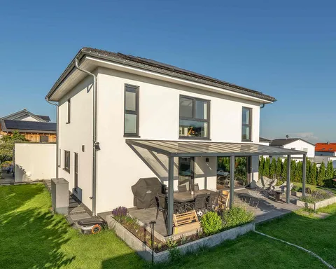 Fertighaus Stadtvilla mit weißer Fassade, dunklem Walmdach, großen Fenstern und überdachter Terrasse im Garten.