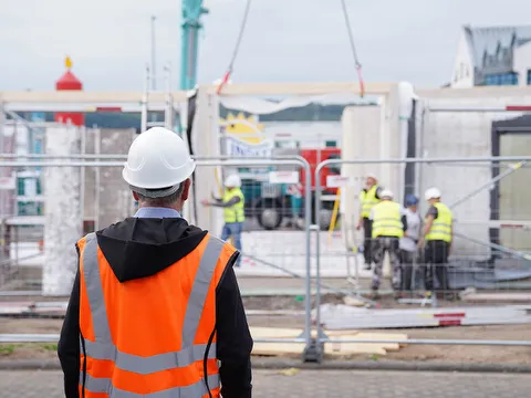 STREIF Baustelle mit Bauarbeiter in Warnweste und Helm vor Fertighauselementen und weiteren Arbeitern bei der Montage
