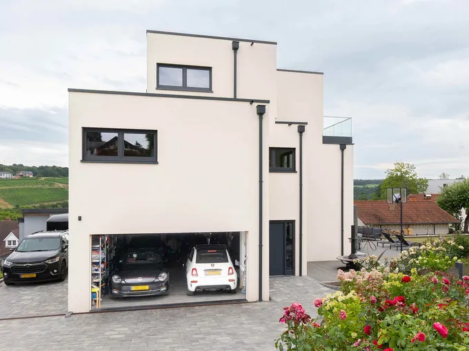 Fertighaus Stadtvilla mit weißer Putzfassade, gestaffeltem Flachdach, dunklen Fensterrahmen und offener Doppelgarage an gepflasterter Einfahrt.