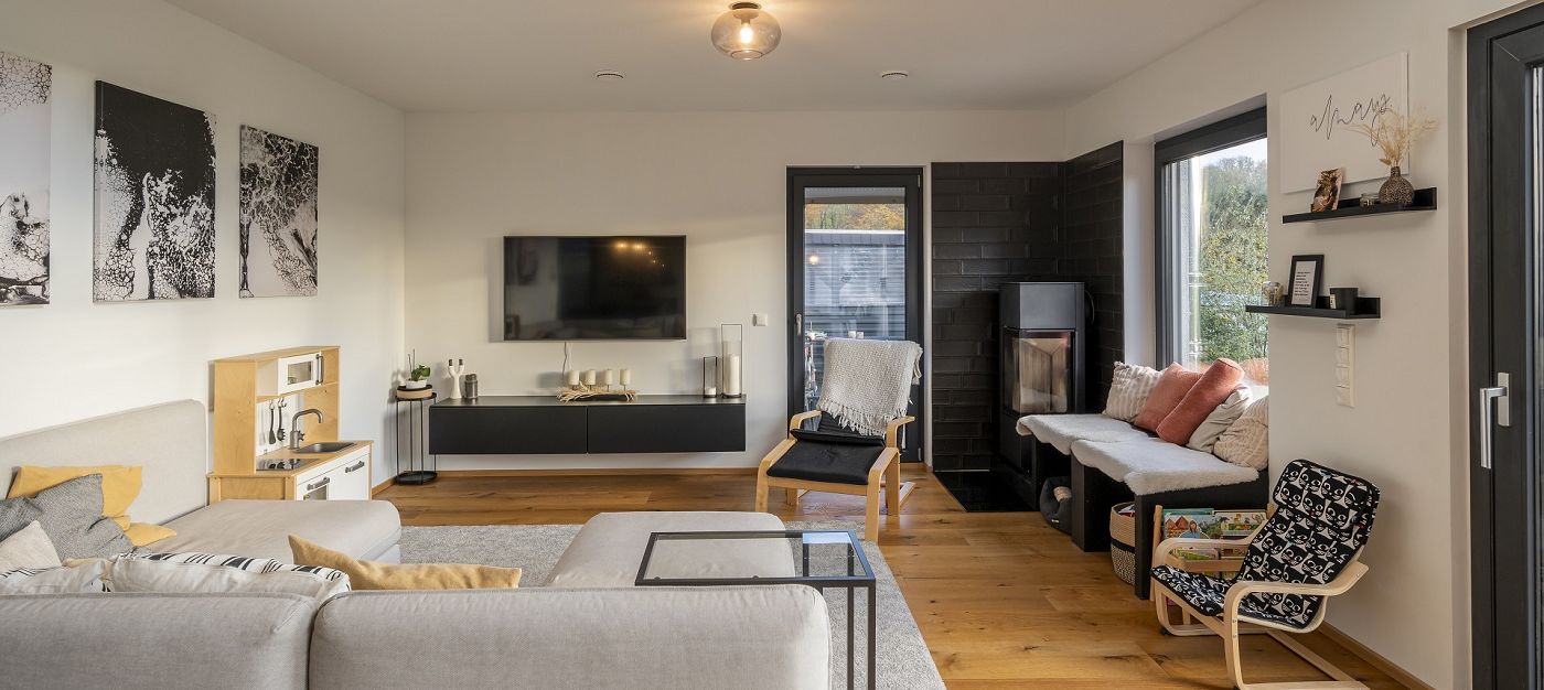 Innenraum Wohnzimmer mit Sofa, Kaminofen, Fernseher, Holzfußboden und großen Fenstern