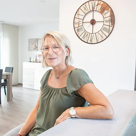 Lächelnde Frau mit Brille sitzt auf einer Couch im hellen modernen Wohnraum, große Wanduhr an der Wand im Hintergrund.