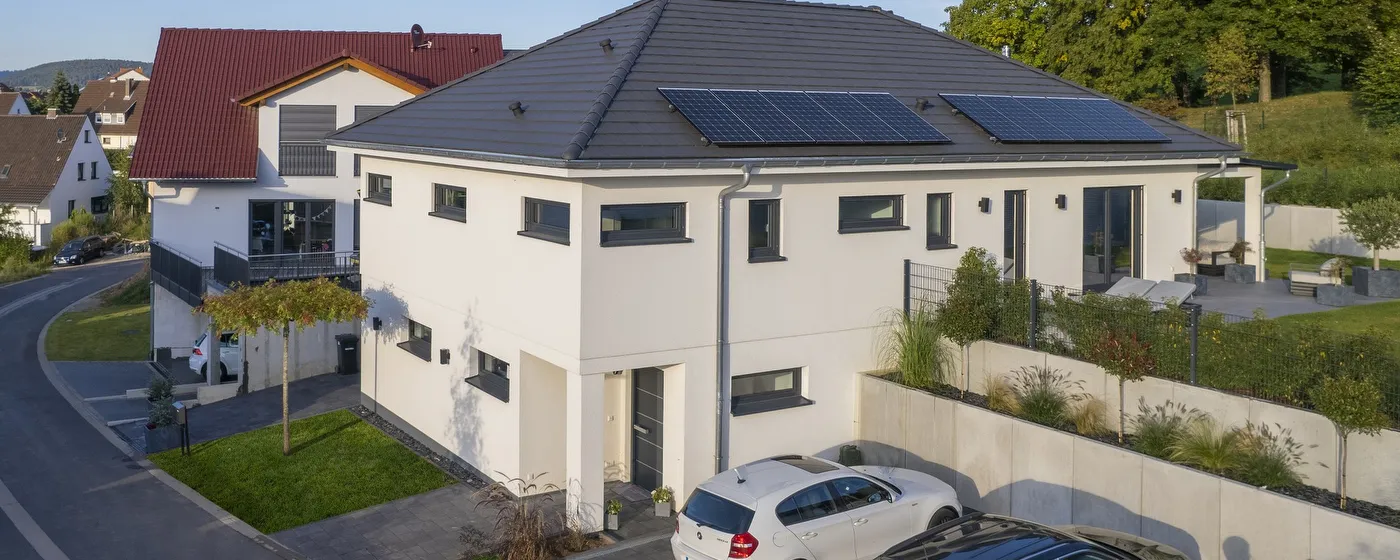 Fertighaus Bungalow, weiße Putzfassade, Walmdach mit Photovoltaik, Außenansicht im Wohngebiet mit Stellplätzen.