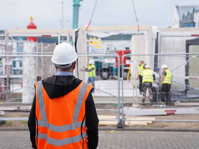 STREIF Baustelle mit Bauarbeiter in Warnweste und Helm vor Fertighauselementen und weiteren Arbeitern bei der Montage