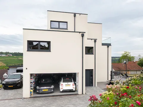 Fertighaus Stadtvilla mit weißer Putzfassade, gestaffeltem Flachdach, dunklen Fensterrahmen und offener Doppelgarage an gepflasterter Einfahrt.