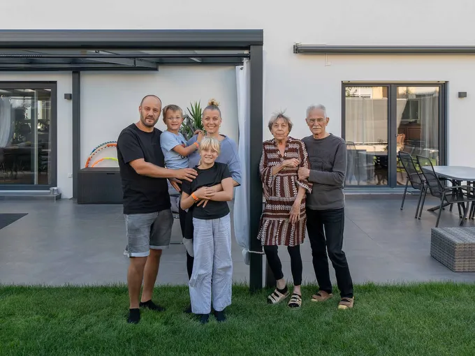 Mehrgenerationenwohnen Familie mit sechs Personen vor modernem Wohnhaus mit Terrasse und Rasenfläche.