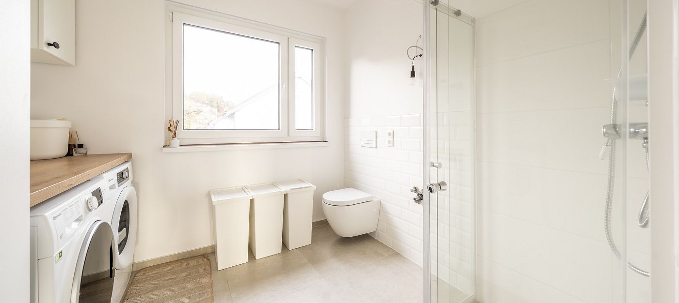 Innenraum Badezimmer mit Dusche, wandhängendem WC, Waschmaschine, Fenster und hellen Fliesen