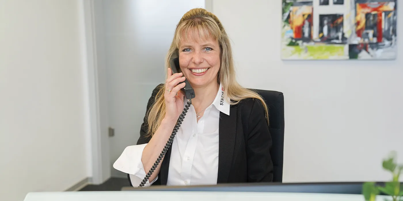 STREIF-Mitarbeiterin mit schwarzem Blazer und weißer Bluse die telefoniert und in die Kamera lächelt.